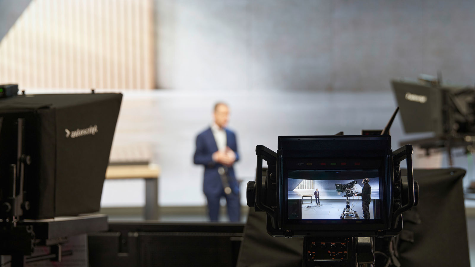 Ein Kameradisplay zeigt eine Aufnahme eines Mannes in einem Anzug, der vor einer minimalistischen Kulisse spricht. Im Vordergrund befinden sich mehrere professionelle Studiokameras und Teleprompter, die die Szene erfassen. Die eigentliche Person im Hintergrund ist unscharf, während die Kamera das scharfe Bild aufnimmt.