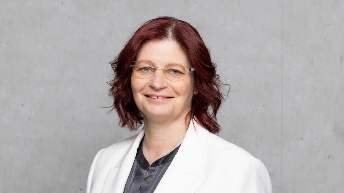 The picture shows Daniela Nowak in front of a gray concrete wall. She is wearing a light-colored blazer, a blouse and glasses.