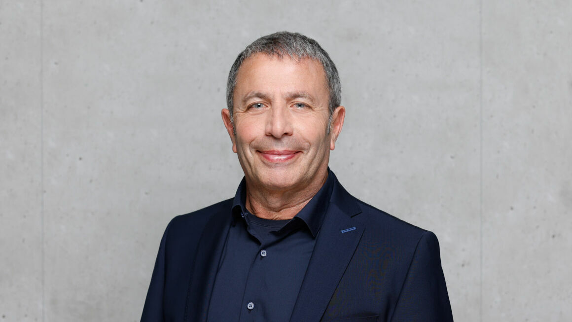 The picture shows Gerardo Scarpino in front of a gray concrete wall. He is wearing a dark suit and a dark shirt.