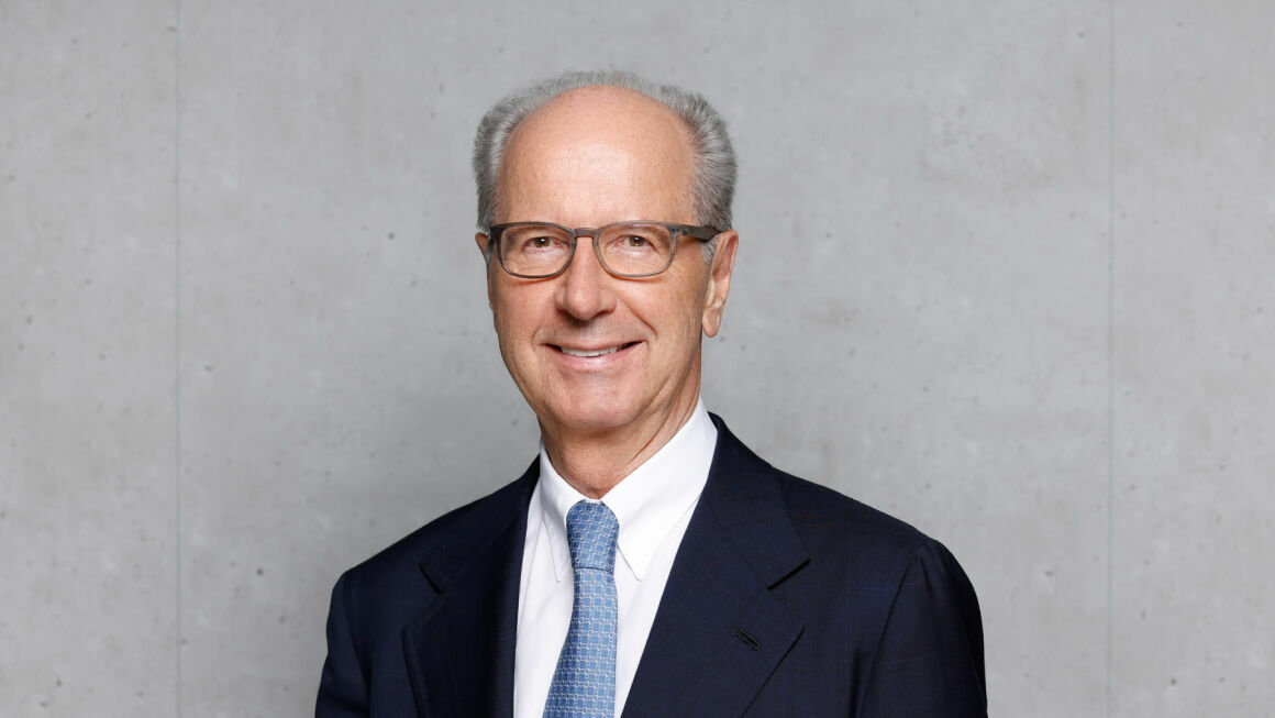 An older gentleman with short gray hair and glasses stands in front of a gray concrete wall. He is wearing a dark blue suit, a white shirt, and a light blue tie. He is smiling warmly at the camera.