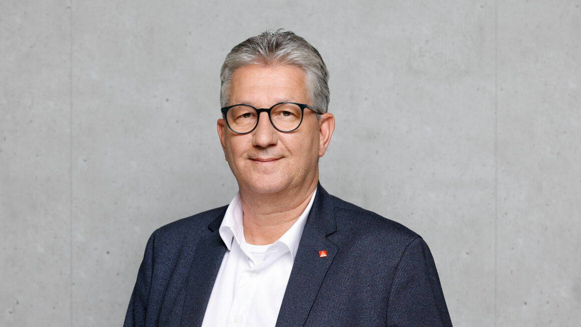 The picture shows Harald Buck in front of a gray concrete wall. He is wearing a suit and a shirt as well as a red pin.