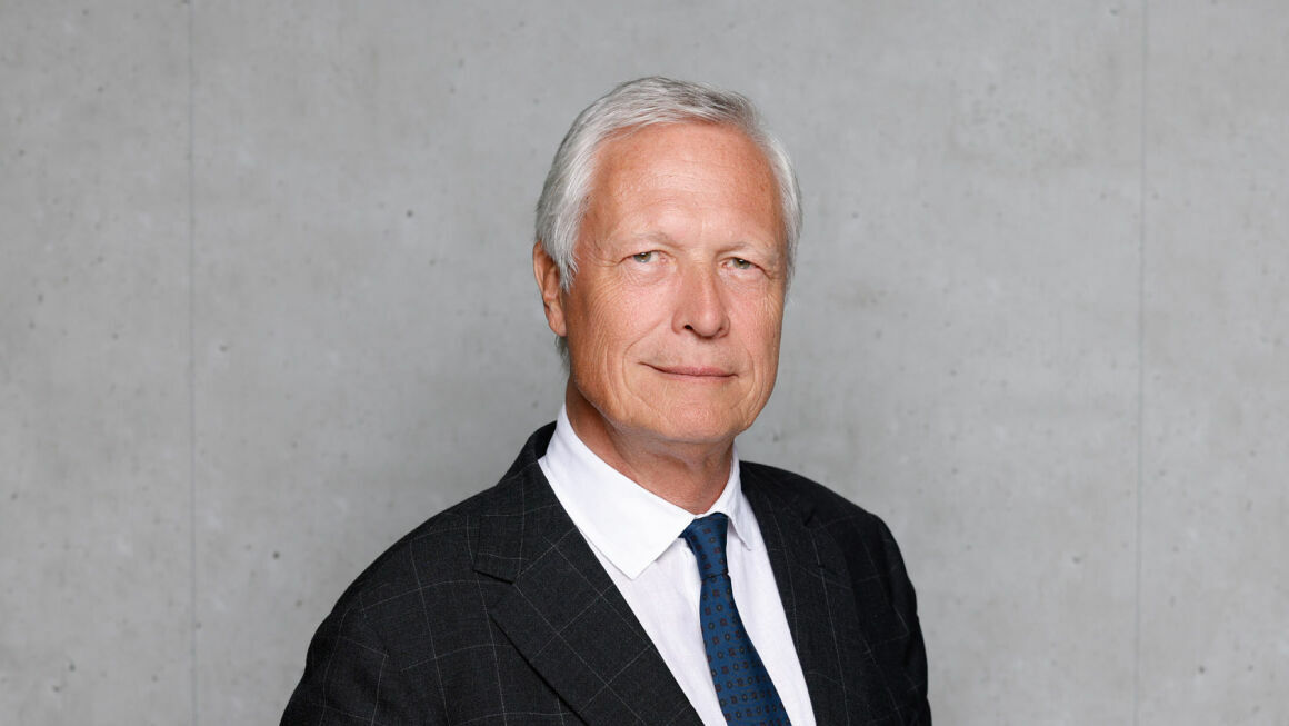 The picture shows Dr. Günther Horvath in front of a grey concrete wall. He is wearing a suit, a tie and a shirt.