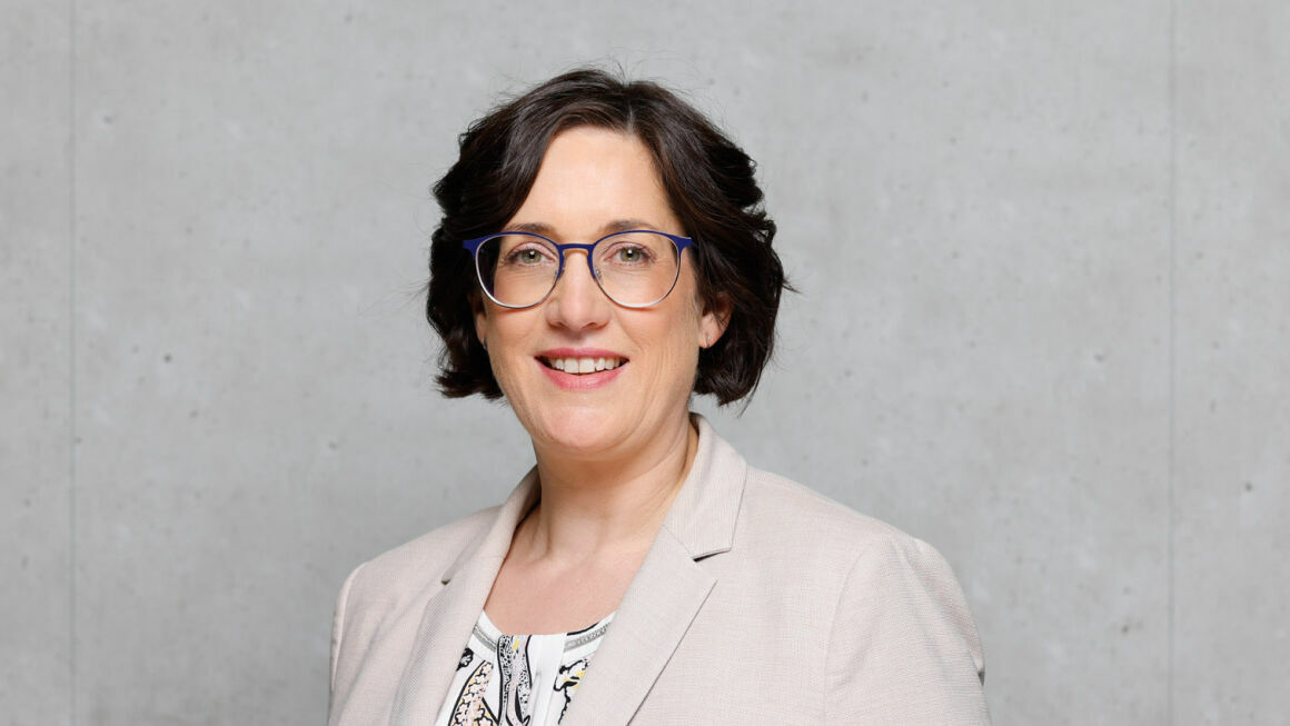 The picture shows Conny Schönhardt in front of a gray concrete wall. She is wearing a light-colored blazer, a blouse and glasses.