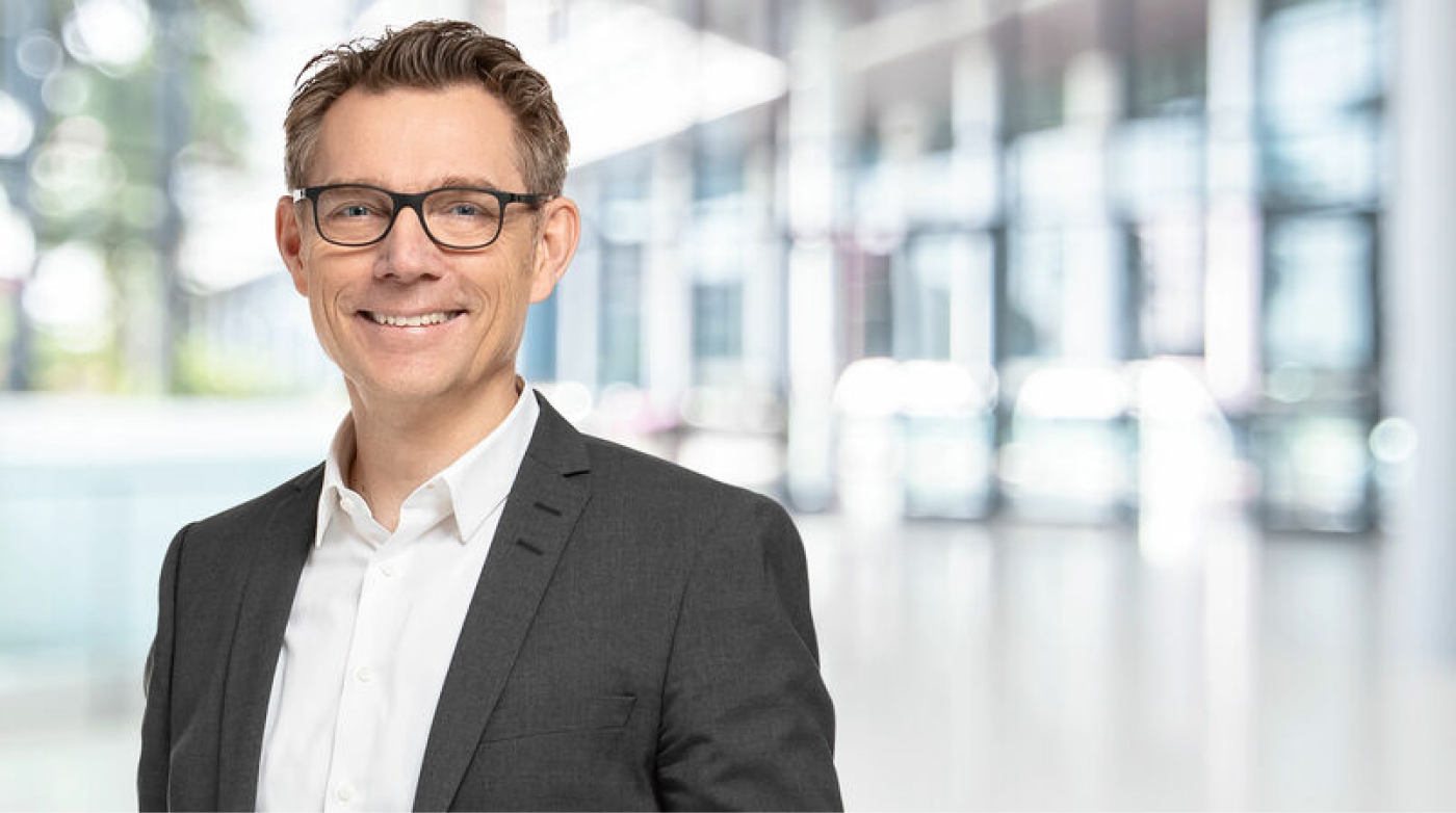 Portrait of a man with short brown hair and glasses, smiling at the camera. He is wearing a dark gray suit with a white shirt. The background features a modern, bright office or conference hall with large glass windows and blurred light reflections.