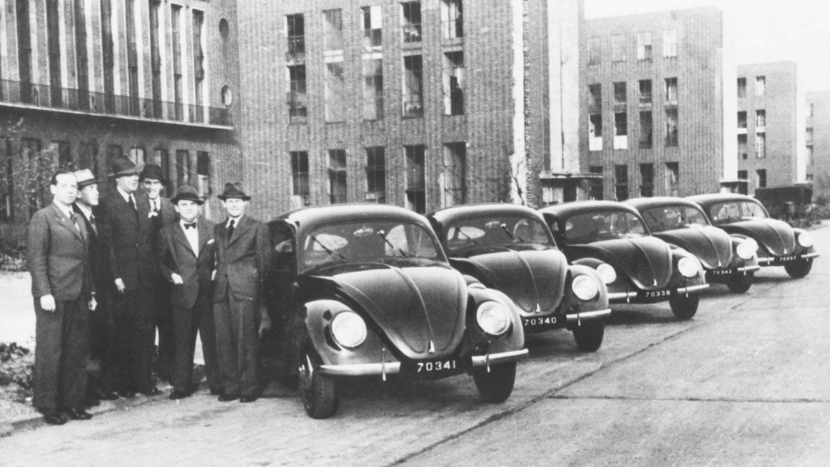 Historische Schwarz-Weiß-Aufnahme einer Gruppe von Männern in Anzügen und Hüten, die neben einer Reihe von Volkswagen Käfern mit aufeinanderfolgenden Nummernschildern stehen. Im Hintergrund sind Backsteingebäude zu sehen.