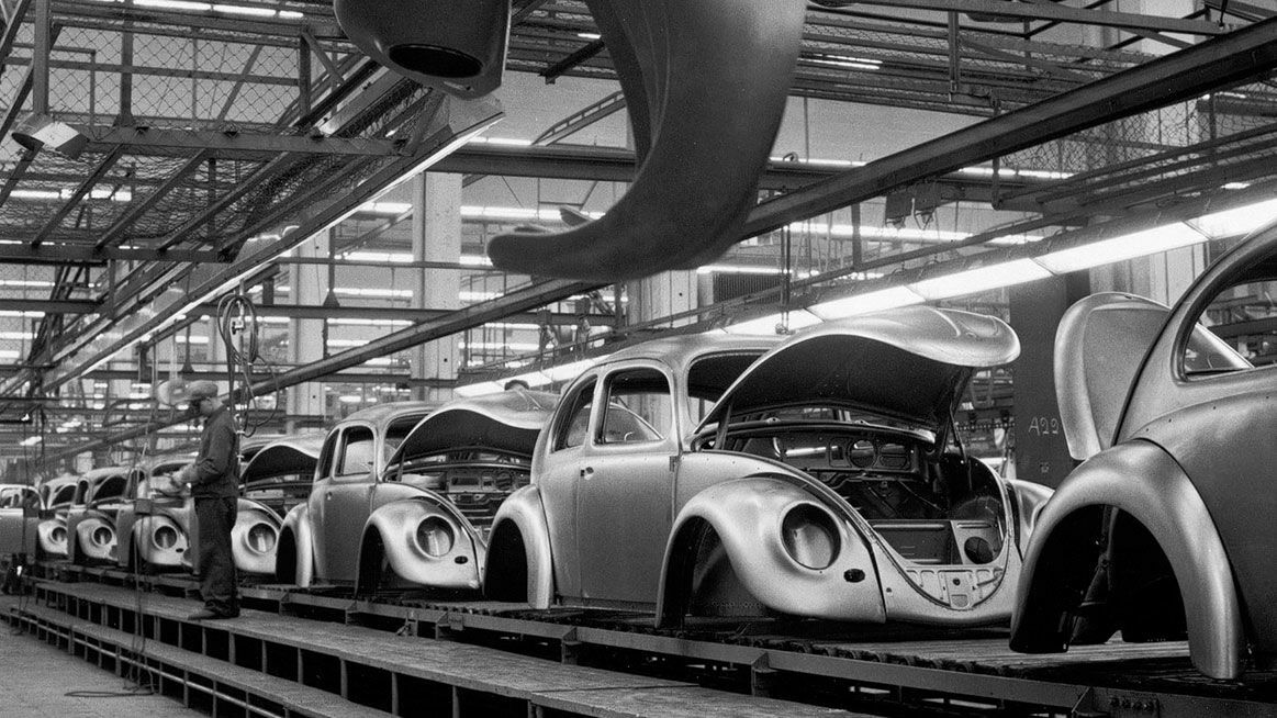 Schwarz-Weiß-Fotografie einer Automobilfertigungslinie, auf der mehrere Volkswagen Käfer mit offenen Motorhauben montiert werden. Ein Arbeiter steht an der Produktionslinie.