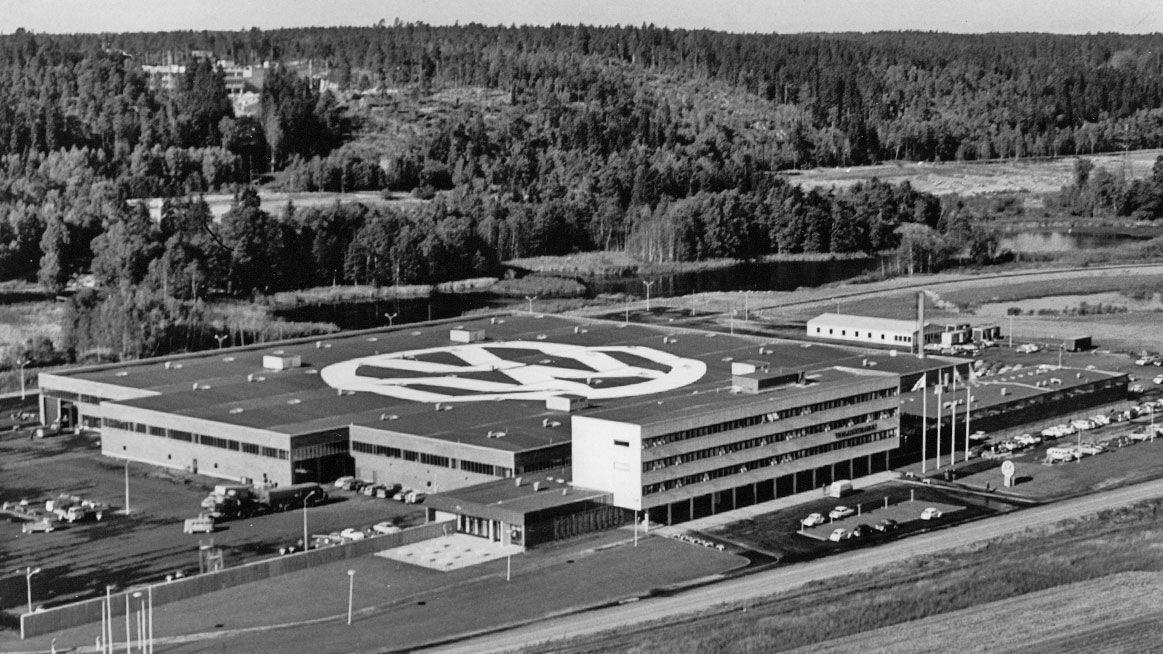 Black-and-white aerial photograph of a Volkswagen plant surrounded by a forested landscape. A large VW logo is visible on the building’s roof. The industrial complex includes parking lots, office buildings, and production halls.