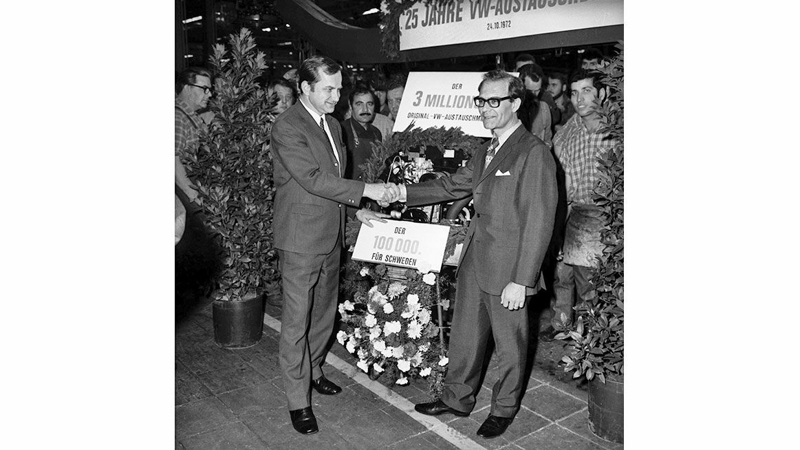Black-and-white photo of two men shaking hands in celebration of the 3-millionth Volkswagen car. In the background, a sign reads “THE 3 MILLIONTH” and “THE 100,000TH FOR SWEDEN.” Flowers and plants are visible in the surroundings.