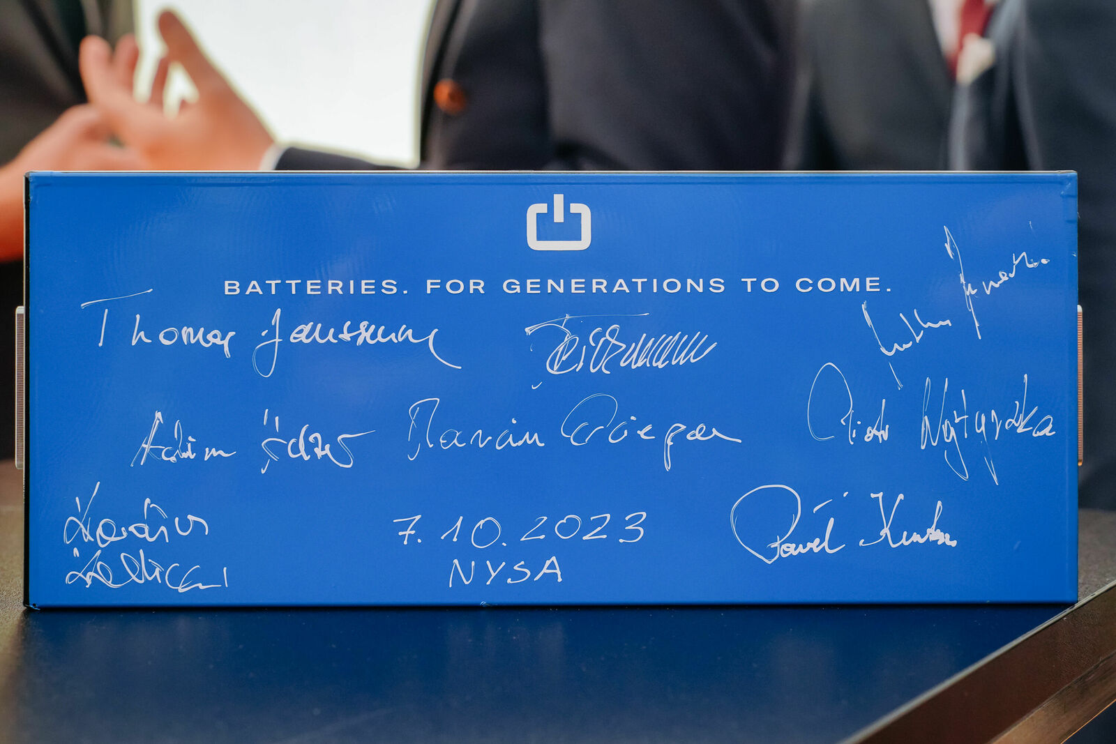 A blue battery with the inscription "BATTERIES. FOR GENERATIONS TO COME." It is signed by multiple individuals, including names like Thomas Jaumann, Marian Górzepa, and Piotr Wojtyczka. The date 7.10.2023 and location Nysa are also written on the battery. In the background, blurred people in suits can be seen.