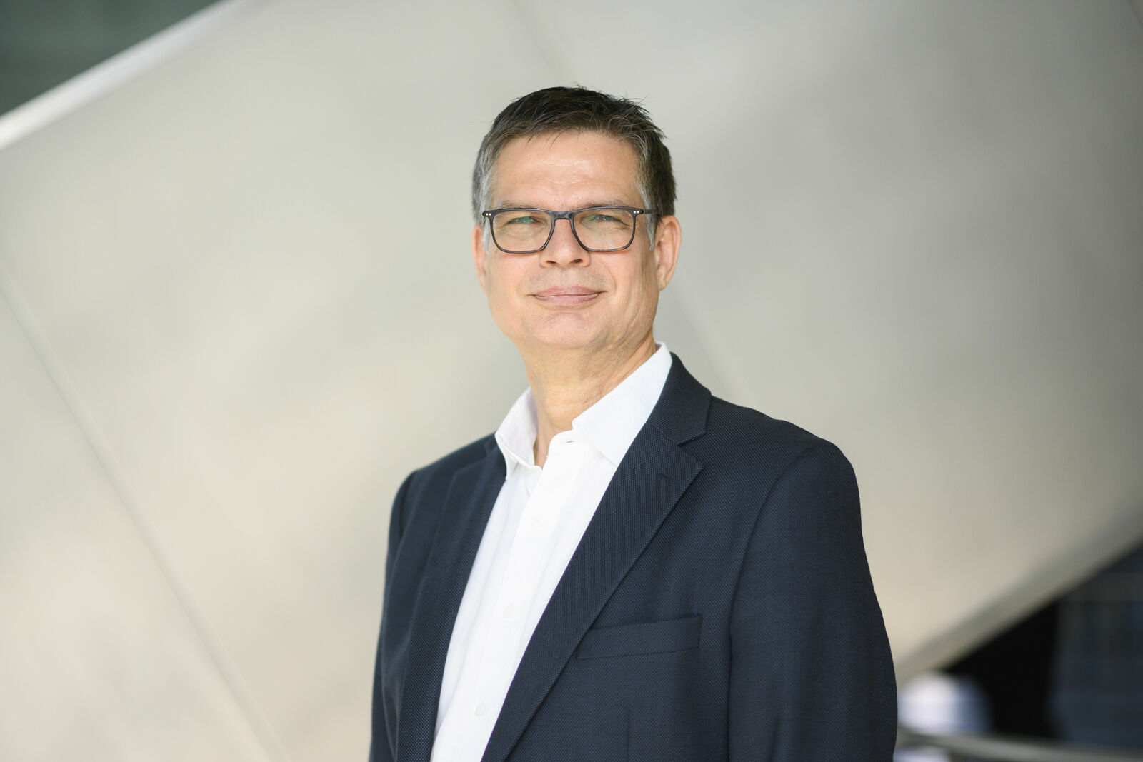 Portrait of a businessman wearing glasses, standing in front of a modern, architecturally interesting background.