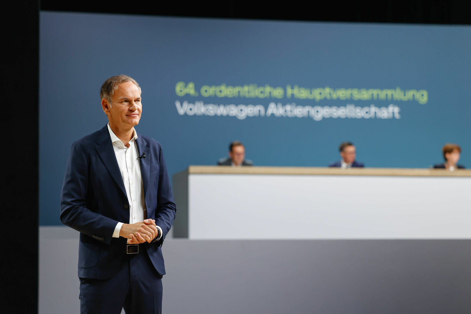 Man in a suit standing at the front of the stage at the 64th ordinary general meeting of Volkswagen Aktiengesellschaft, with other people seated at the podium in the background.