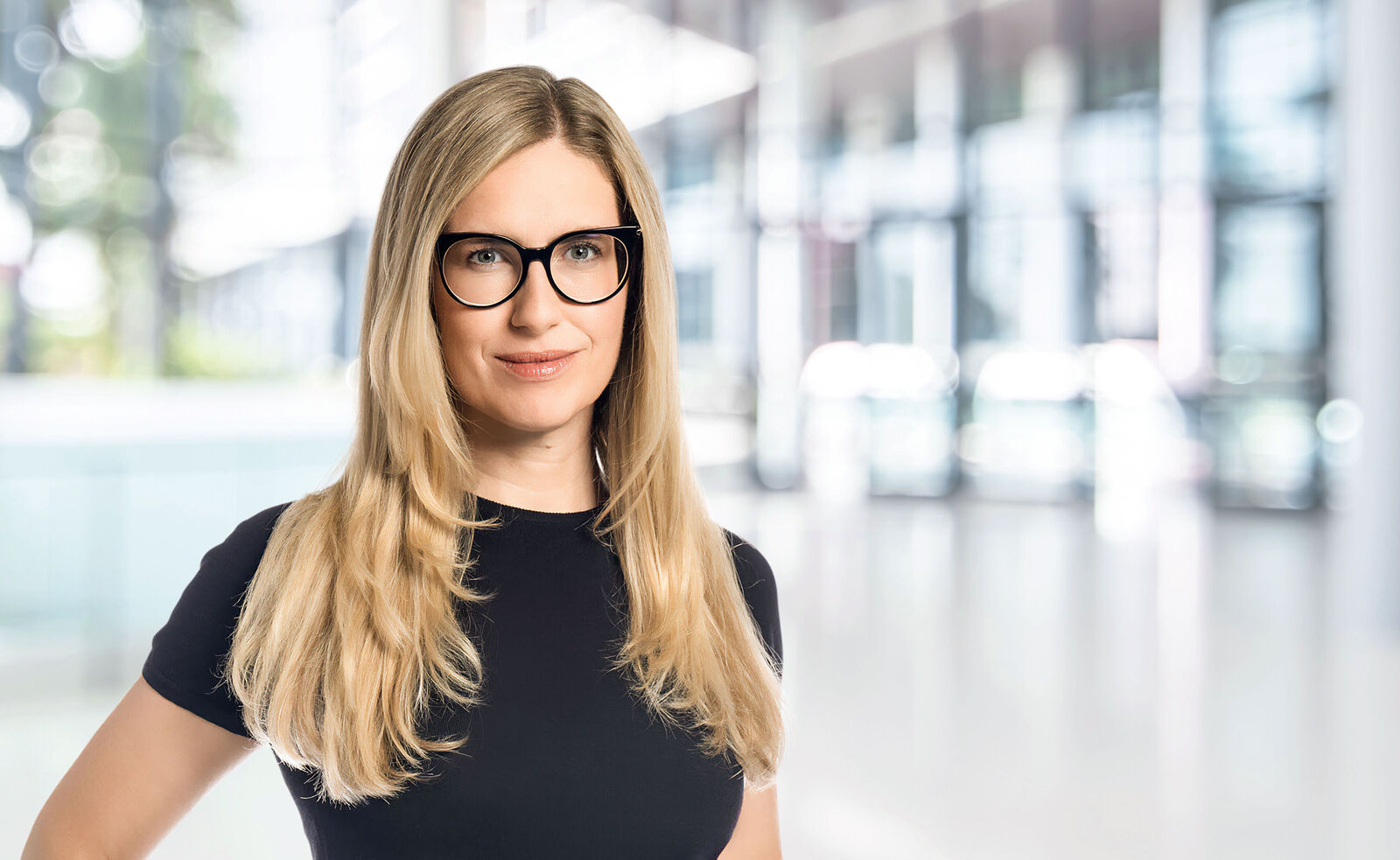 Portrait of a woman with long blonde hair and black glasses, wearing a black top. She looks directly at the camera with a slight smile. The background is modern and brightly lit.